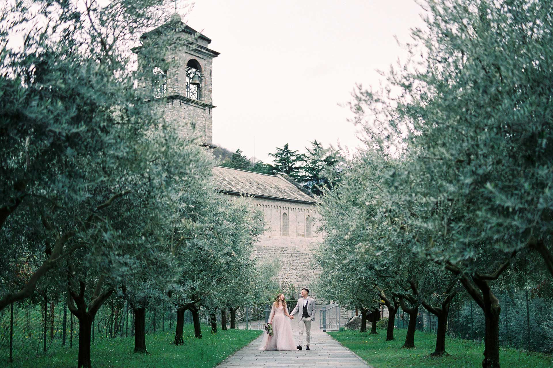 "Pareja caminando de la mano en un jardín rodeado de olivos con una antigua iglesia de piedra al fondo, durante una boda en un entorno natural y romántico.