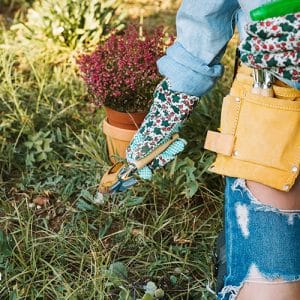 Persona con guantes de jardinería estampados y cinturón de herramientas cuidando su jardín. Sostiene unas tijeras de poda mientras trabaja en el césped. Cuidar Tu Jardín con dedicación y herramientas adecuadas.